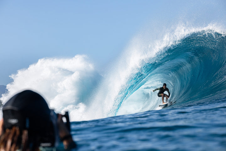 A wetsuit-clad surfer crouches inside a massive turquoise barrel wave while a water photographer shoots from the foreground on a bright blue-sky day.